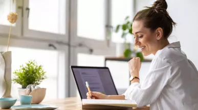 Shows a woman writing in her notepad next to her laptop